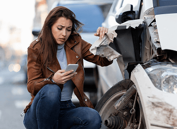 Woman kneels beside her damaged car and looks at her phone worried