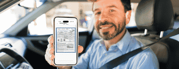 A man in a driver's seat holding a smartphone that displays a PDF of the Banjercito Temporary Vehicle Importation Permit TIP. Best Mexico Insurance