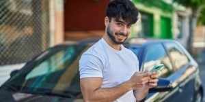 Young Hispanic man using WhatsApp on a smartphone leaning on a car in Mexico. Cheap Mexican Auto Insurance